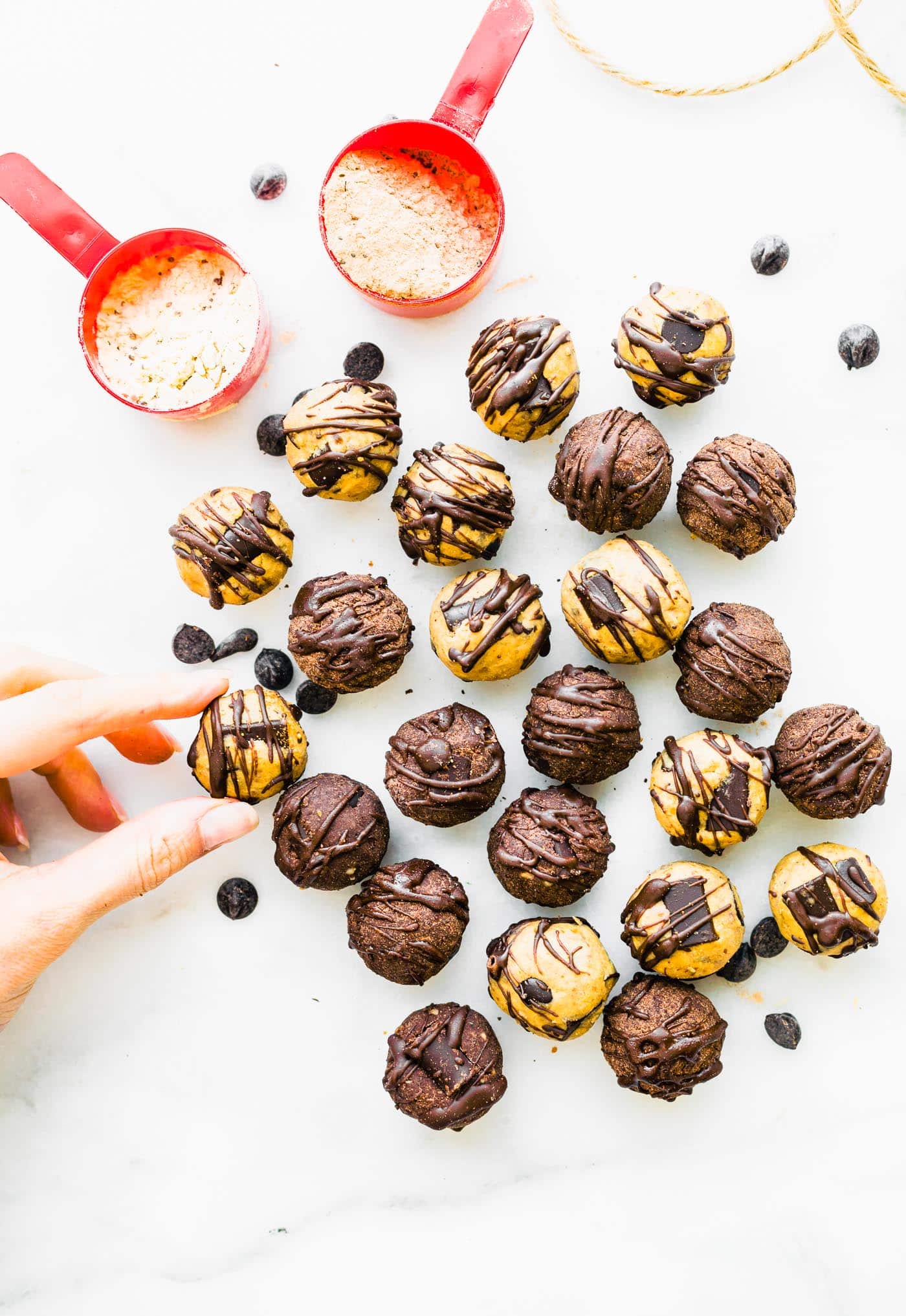 Several dark chocolate protein balls with chocolate drizzle on white counter, one ball being picked up.