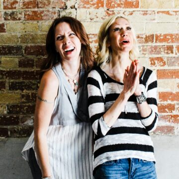 Two women leaning against each other against brick wall, laughing.