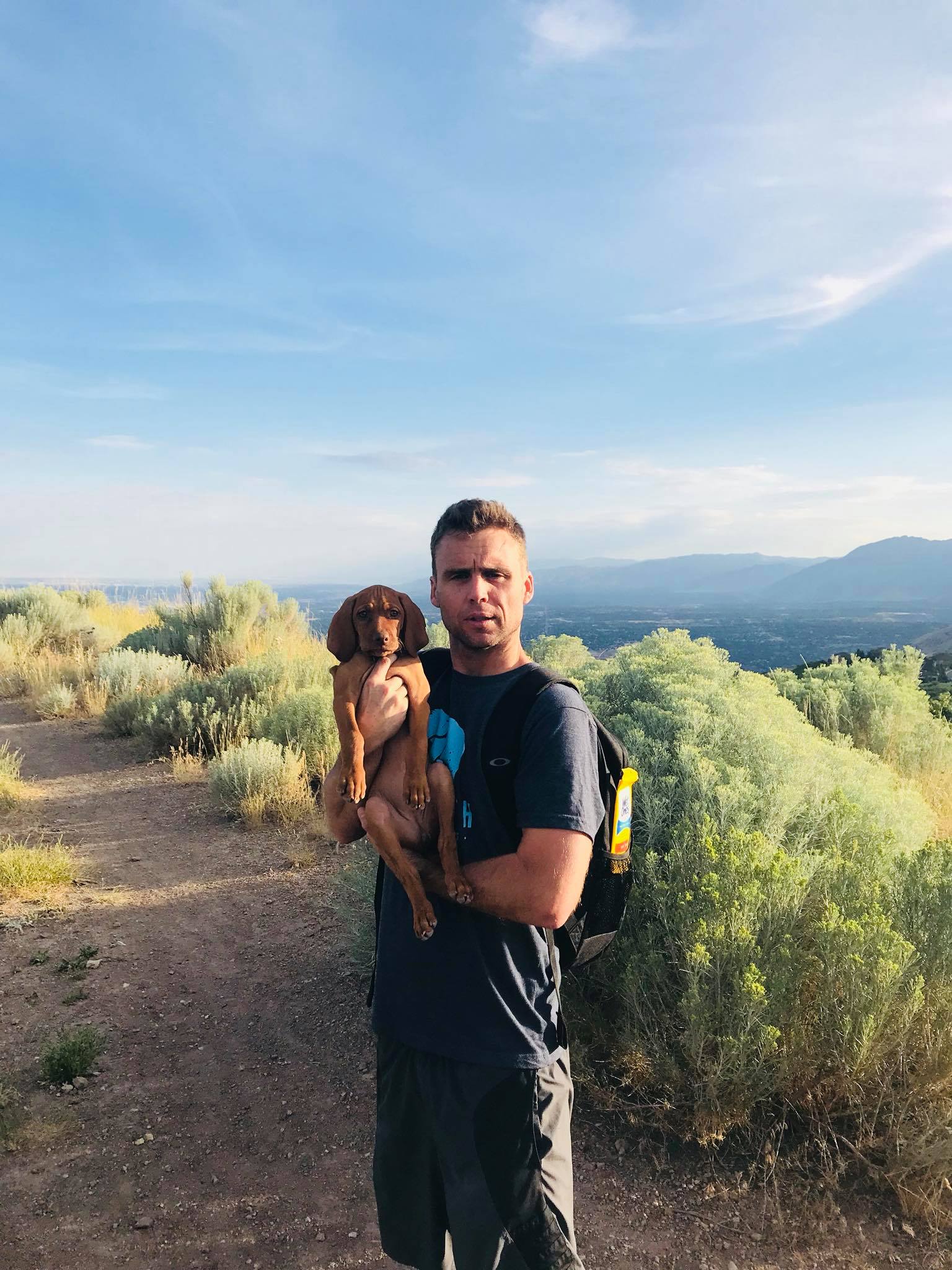 A man wearing a backpack holding a brown vizsla puppy on a trail with mountains in background.