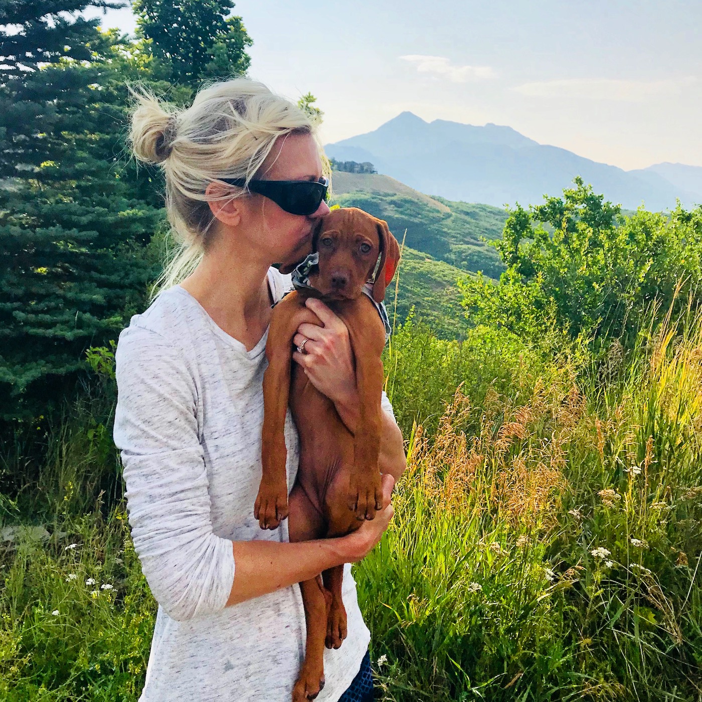 A woman holding a vizsla puppy with green mountainous background.
