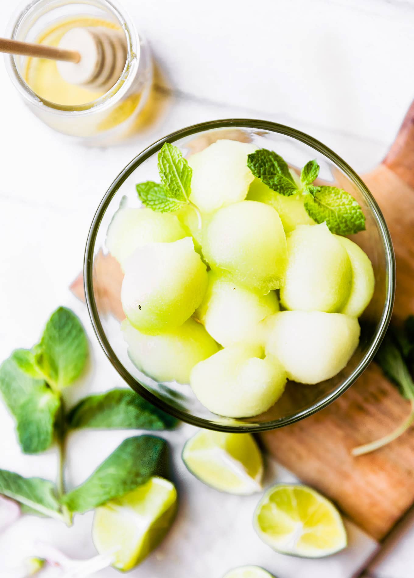 Overhead view melon balls in white glass on wooden cutting board, fresh mint nearby.