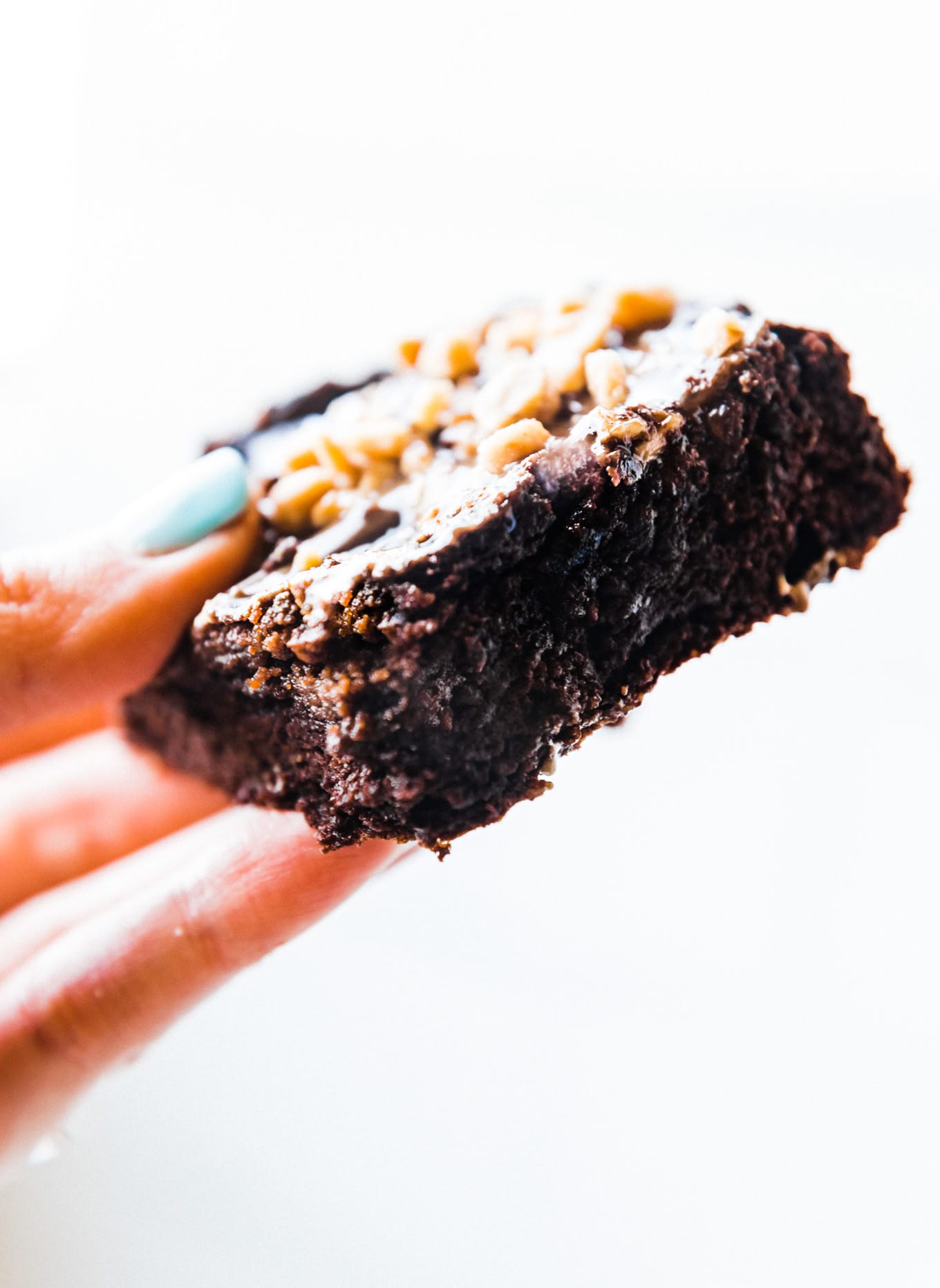 A square piece of low carb brownie with chocolate tahini topping and crushed nuts being held up against white background.
