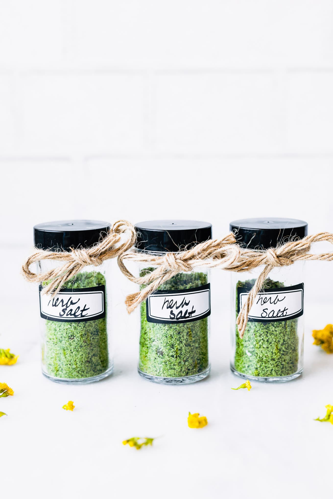 Three clear jars filled with herb salt, black lids, small twine bow tied to each lid.