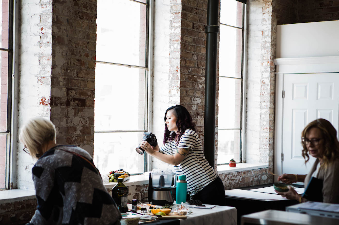 Tables set up by tall windows, filled with food photography layouts, women with cameras photographing food.