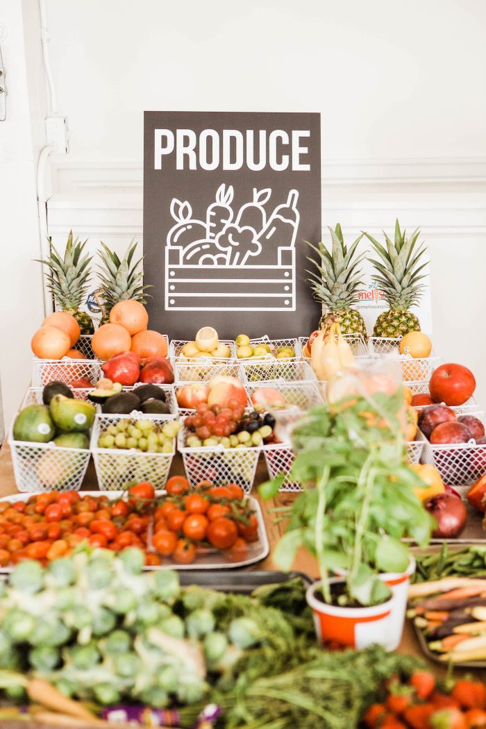 Black sign with white words and graphics for 'produce', fresh produce in white baskets in front of sign.
