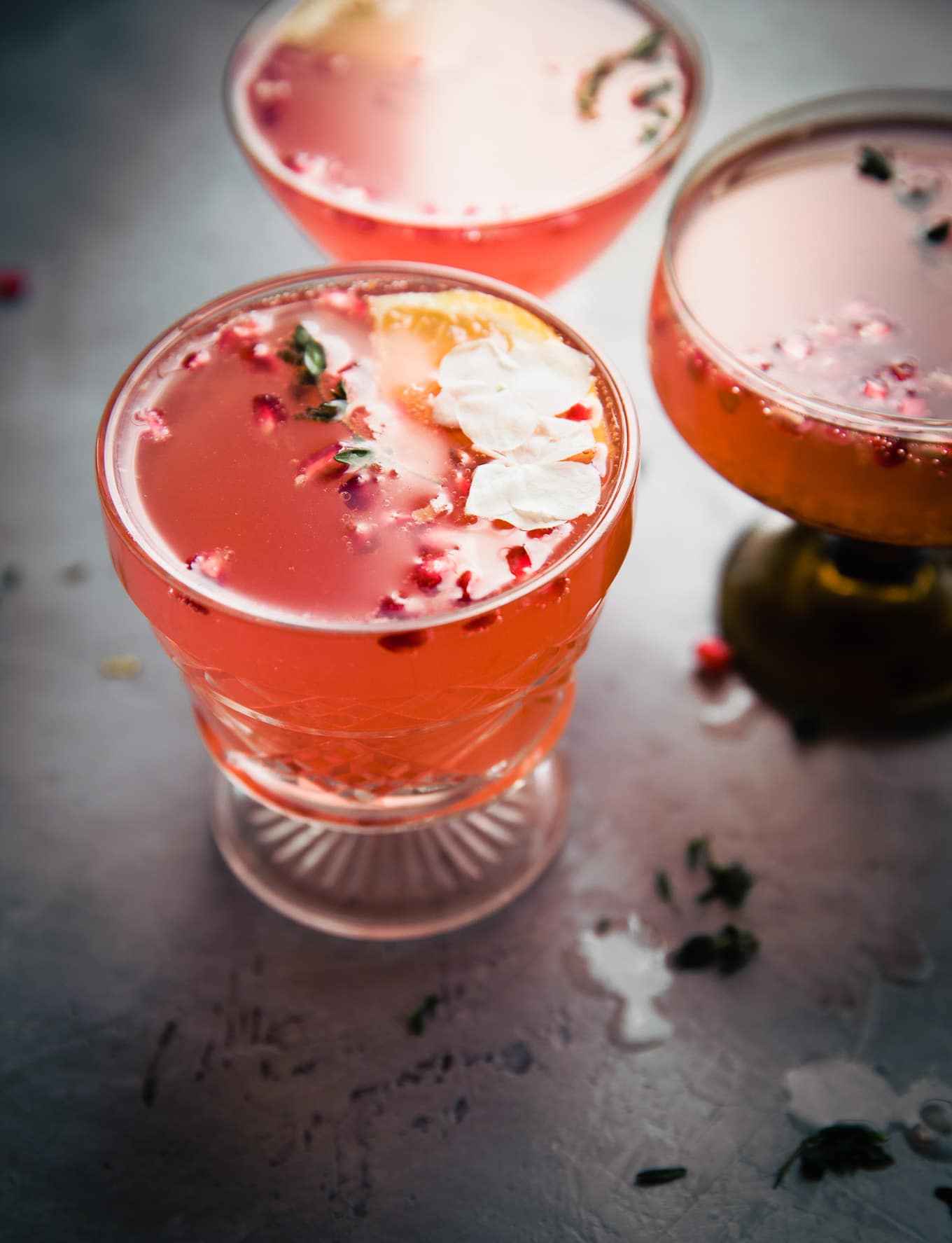 Three drinks with floating pomegranate arils, orange slices, and flowers on dark background.