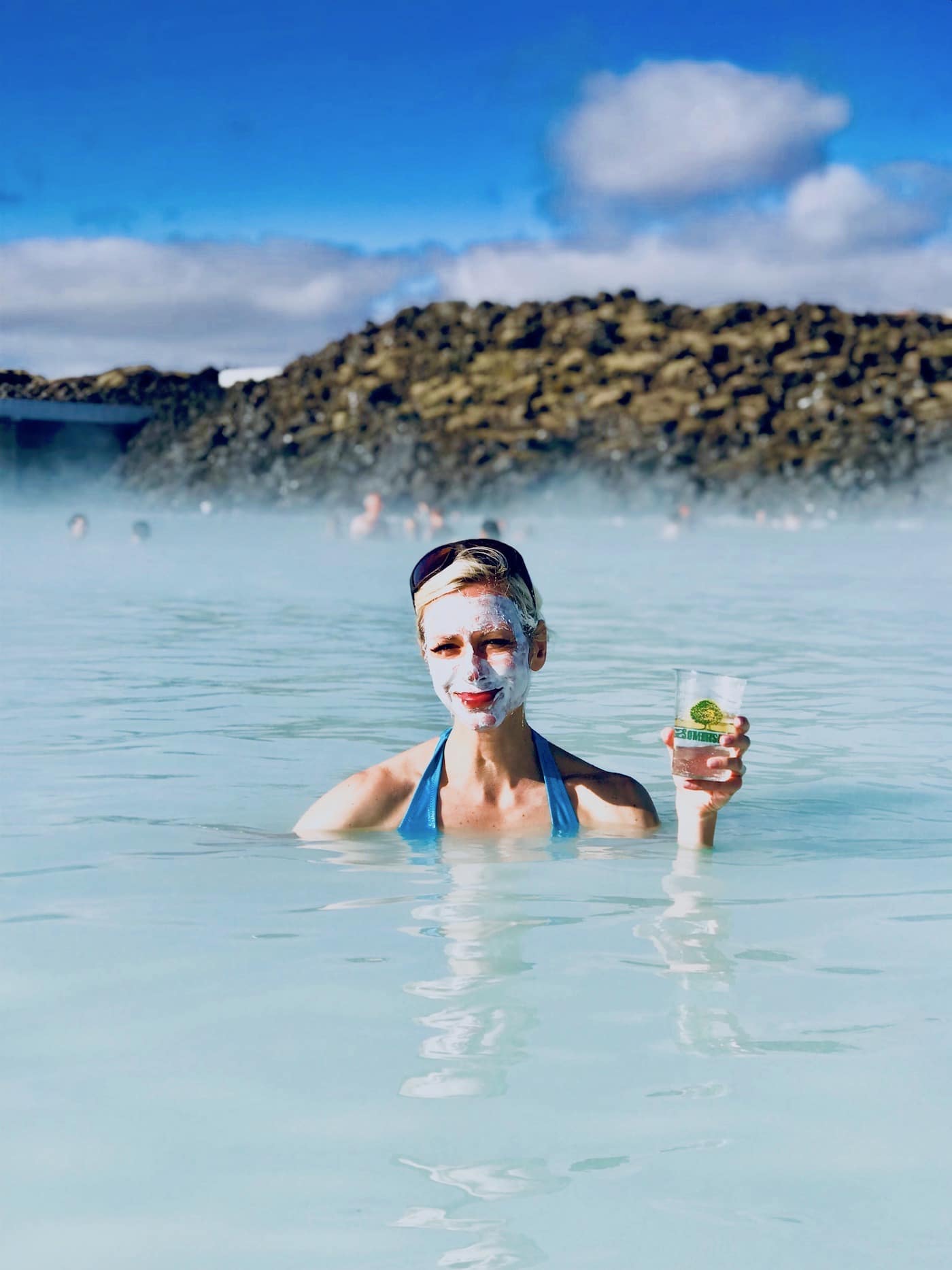 A woman with a white face mud mask holding a drink floating in water.