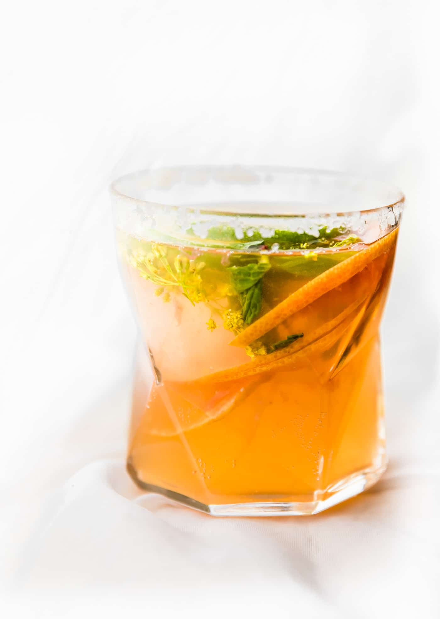 A glass filled with citrus kombucha mezcal cocktail topped with orange slices and herbs.