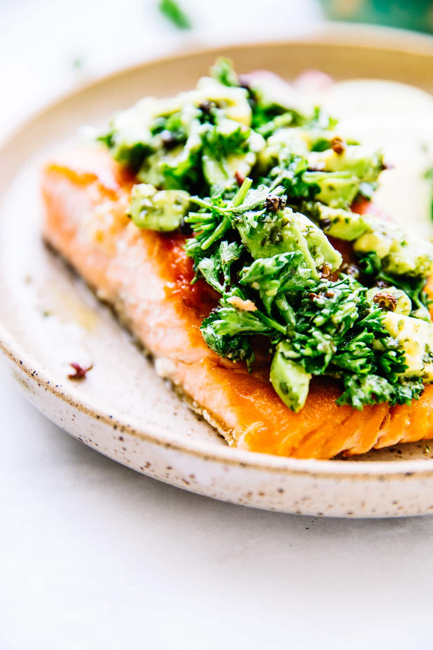 Close up view seared salmon fillet topped with avocado gremolata served in a shallow speckled bowl.