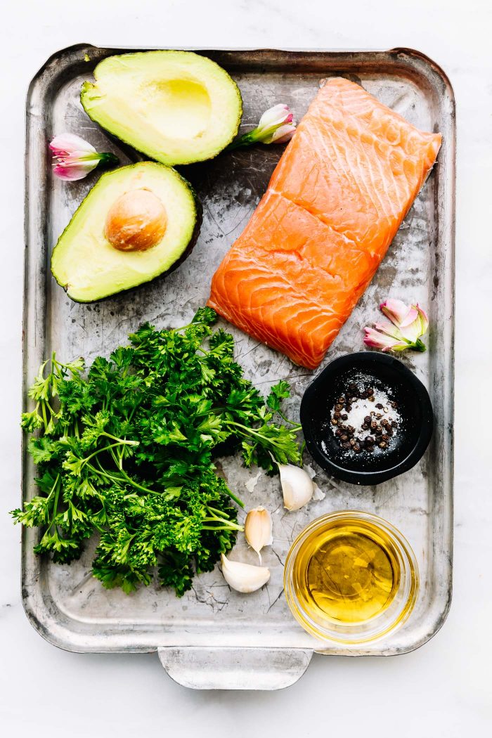 Silver baking sheet with salmon fillet, half an avocado, fresh herbs, and small jar of oil.