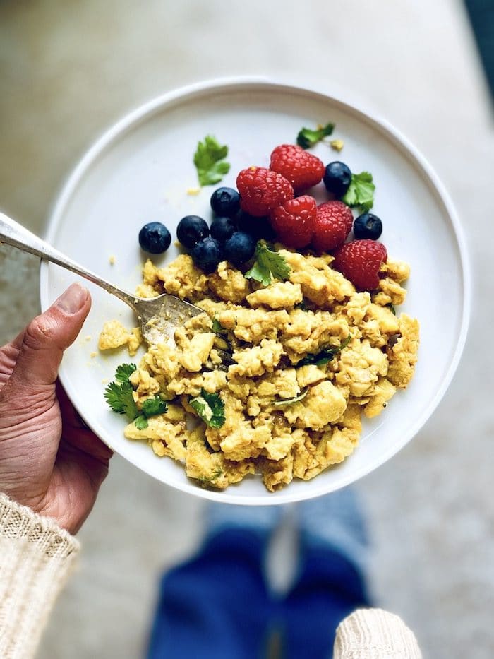 A hand holding a white plate filled with scrambled eggs and fresh berries.