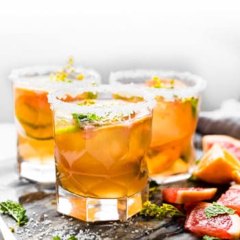 Three glasses of kombucha mezcal topped with orange slices and herbs.