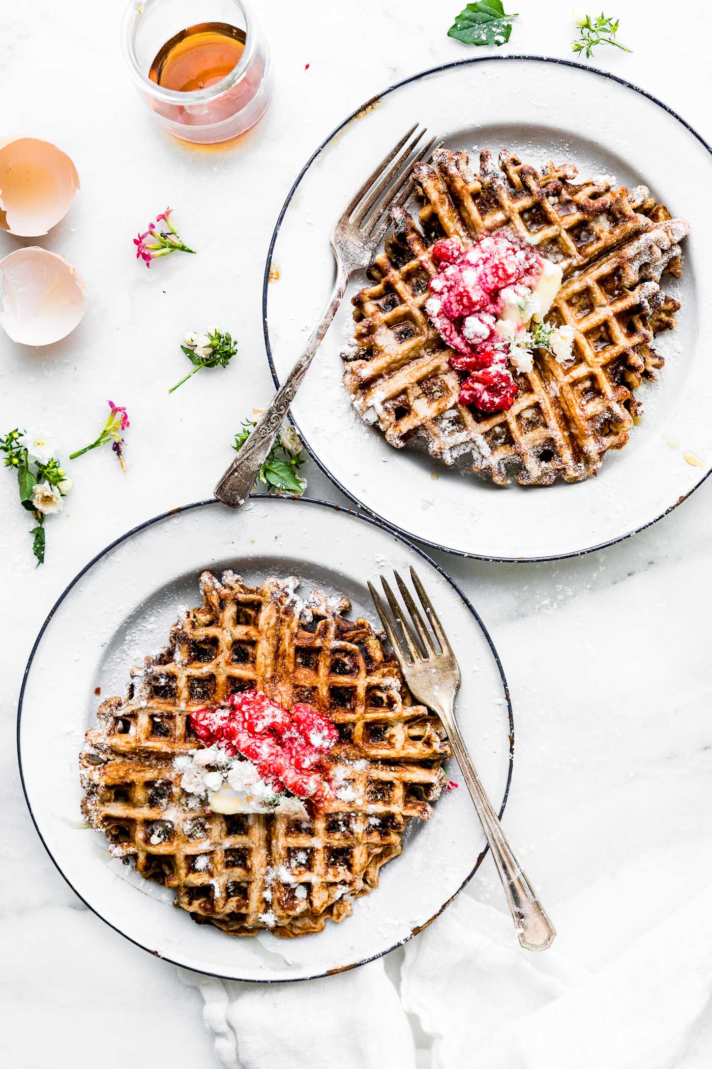Paleo waffles, golden brown, on white plates topped with berries, butter, and maple syrup.
