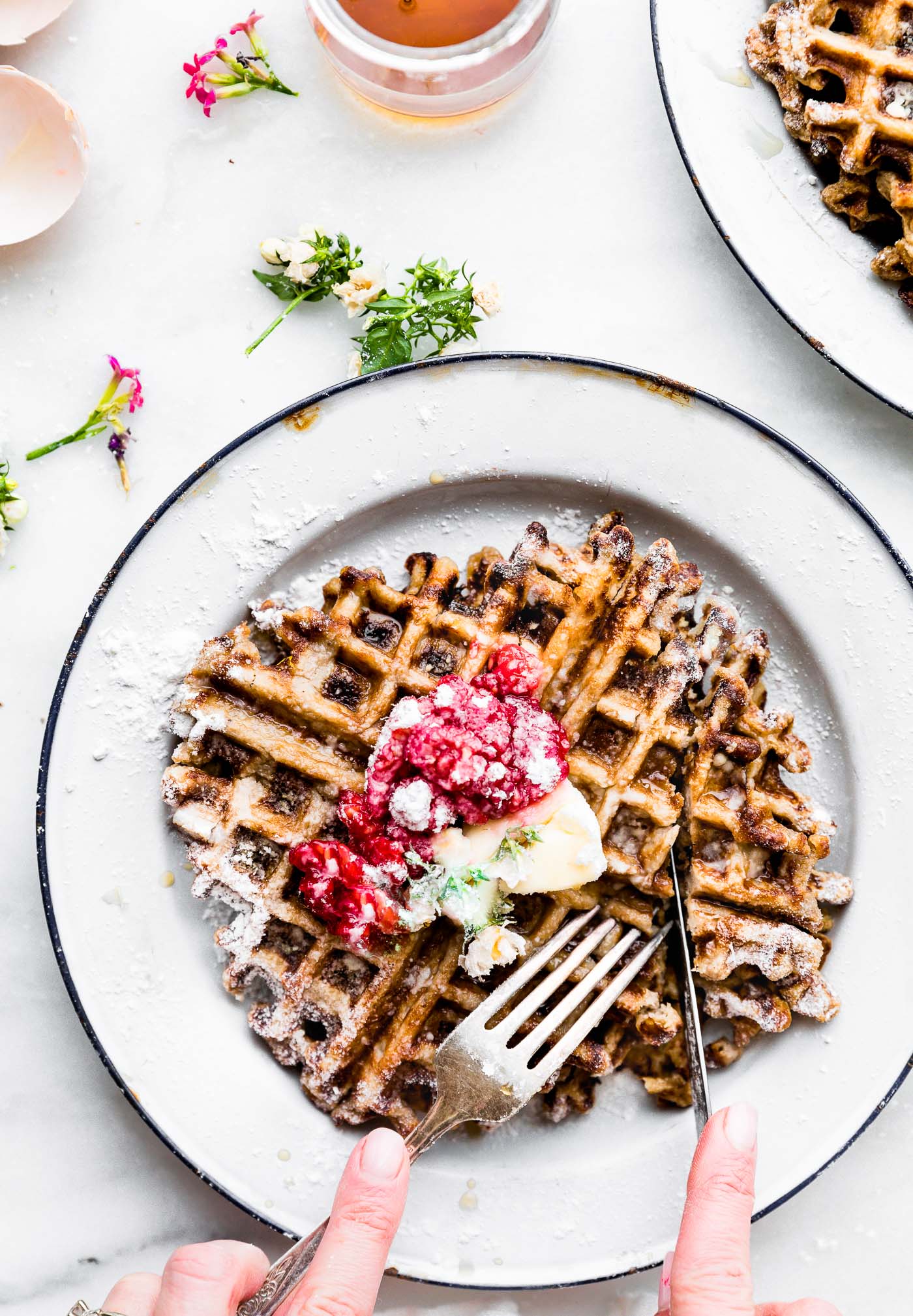Close up view Paleo waffles on white plate topped with mashed berries, butter and powdered sugar.