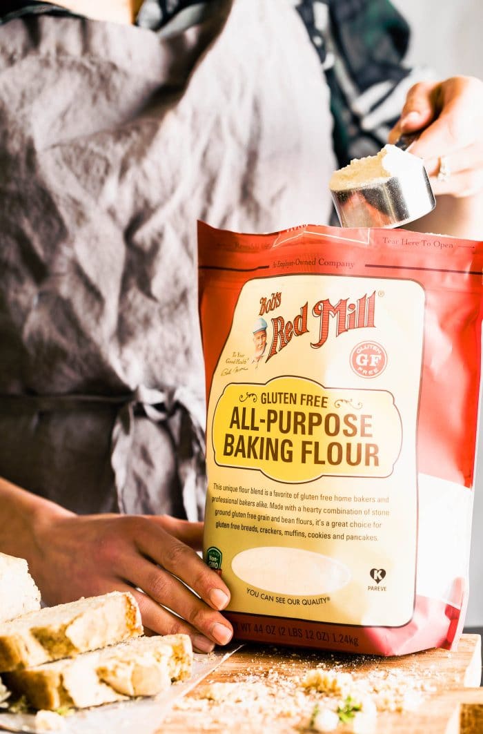 A woman scooping out a measuring cup full of all purpose baking flour from a red Bob's Red Mill bag.