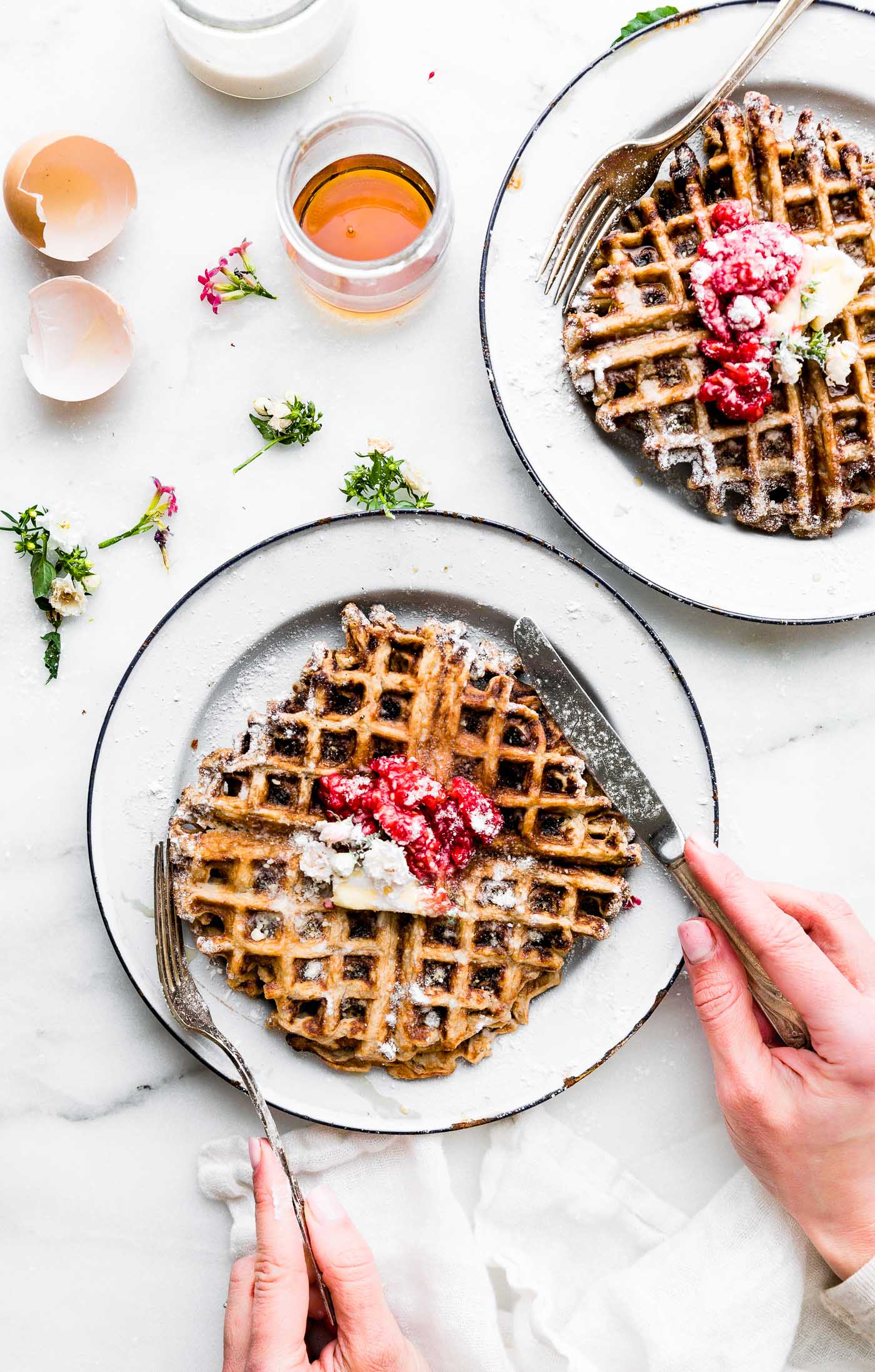 Two white plates with stacks of Paleo waffles, topped with berries, butter, maple syrup, and powdered sugar.