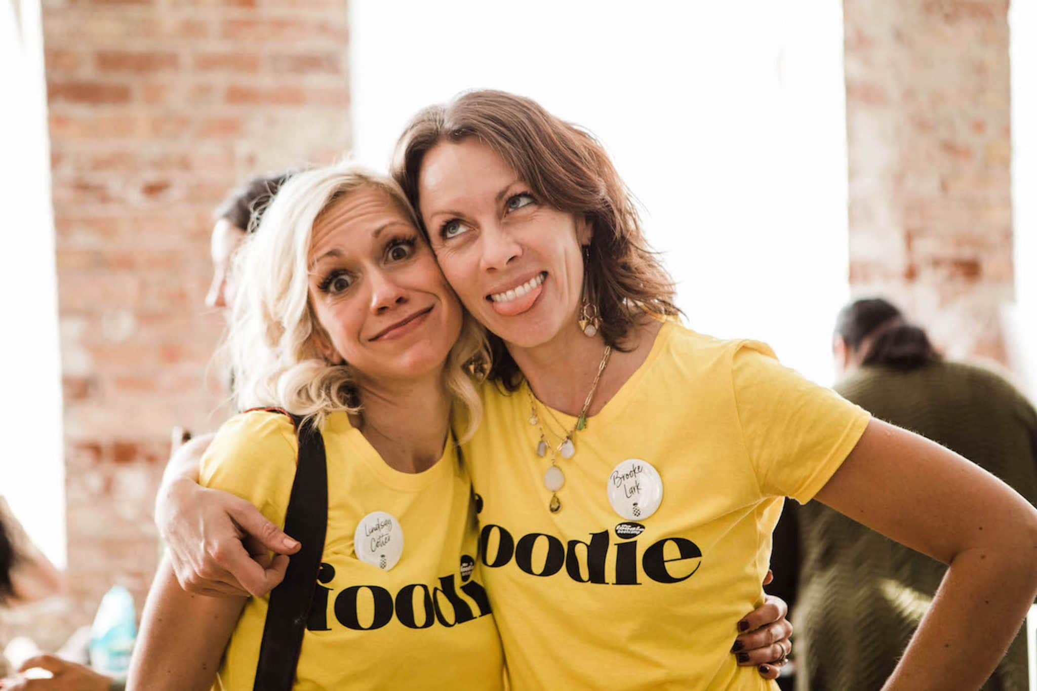 Two woman wearing yellow shirts with 'foodie' screen printed over top, hugging each other.