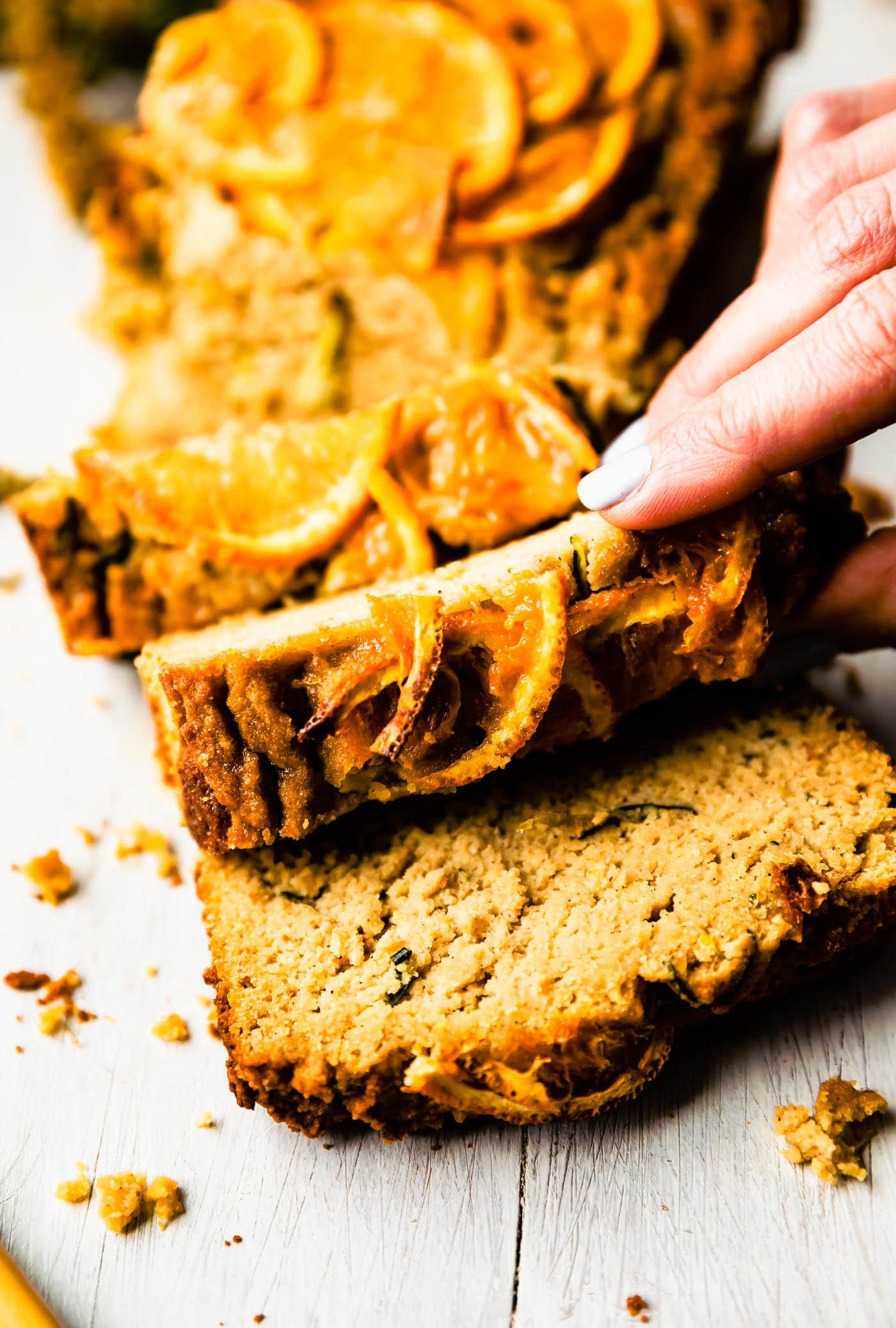 A hand lifting a thick slice of orange zucchini bread from a cutting board holding several slices.