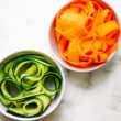 Two small white bowls filled with zucchini noodles and carrot noodles.