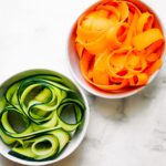 Two small white bowls filled with zucchini noodles and carrot noodles.
