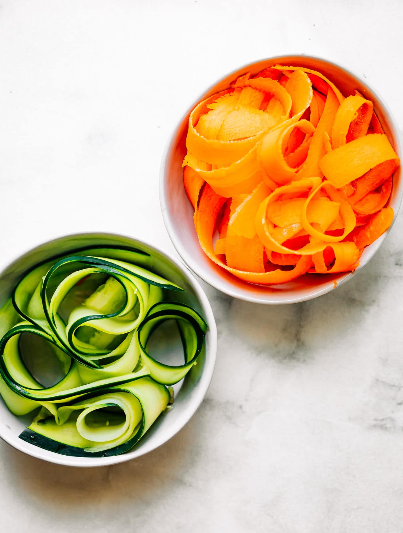 Small white bowl filled with carrot peels and white bowl filled with cucumber strips for veggie noodles.