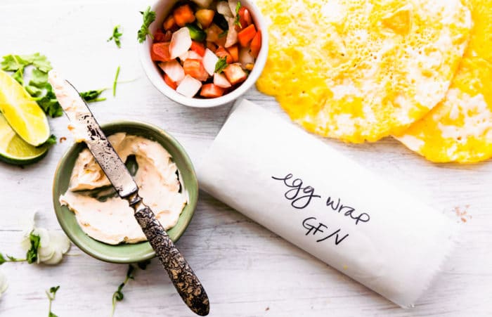 An egg wrap rolled in parchment paper with labeling on white cutting board with egg wrap ingredients scattered around.