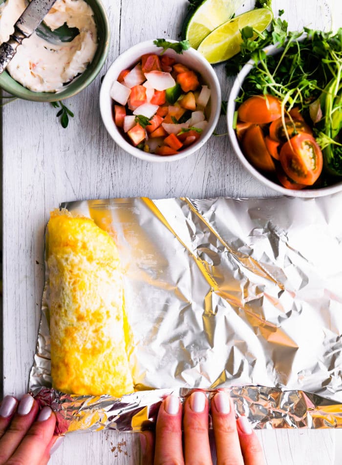 A stuffed egg wrap being rolled in tin foil, on white cutting board with bowls of fresh produce on top.