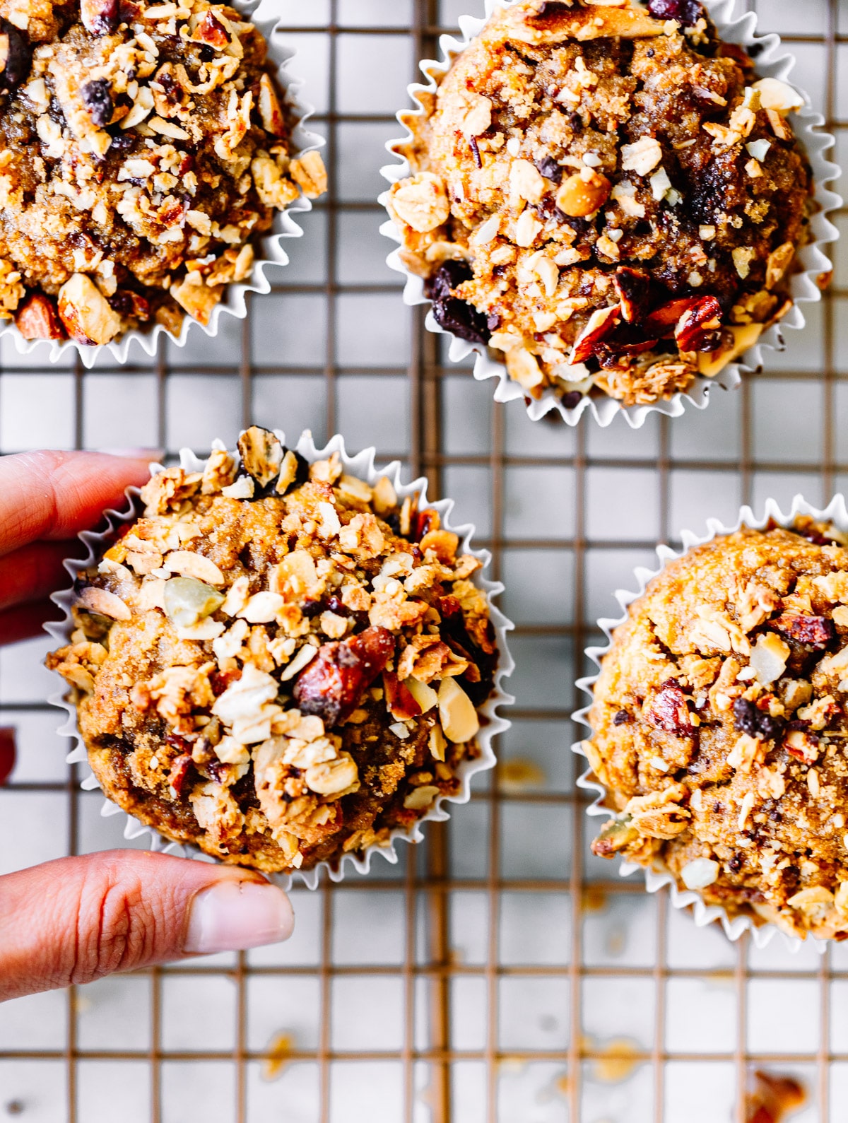 A hand picking up a breakfast muffin topped with muesli from wire cooling rack.