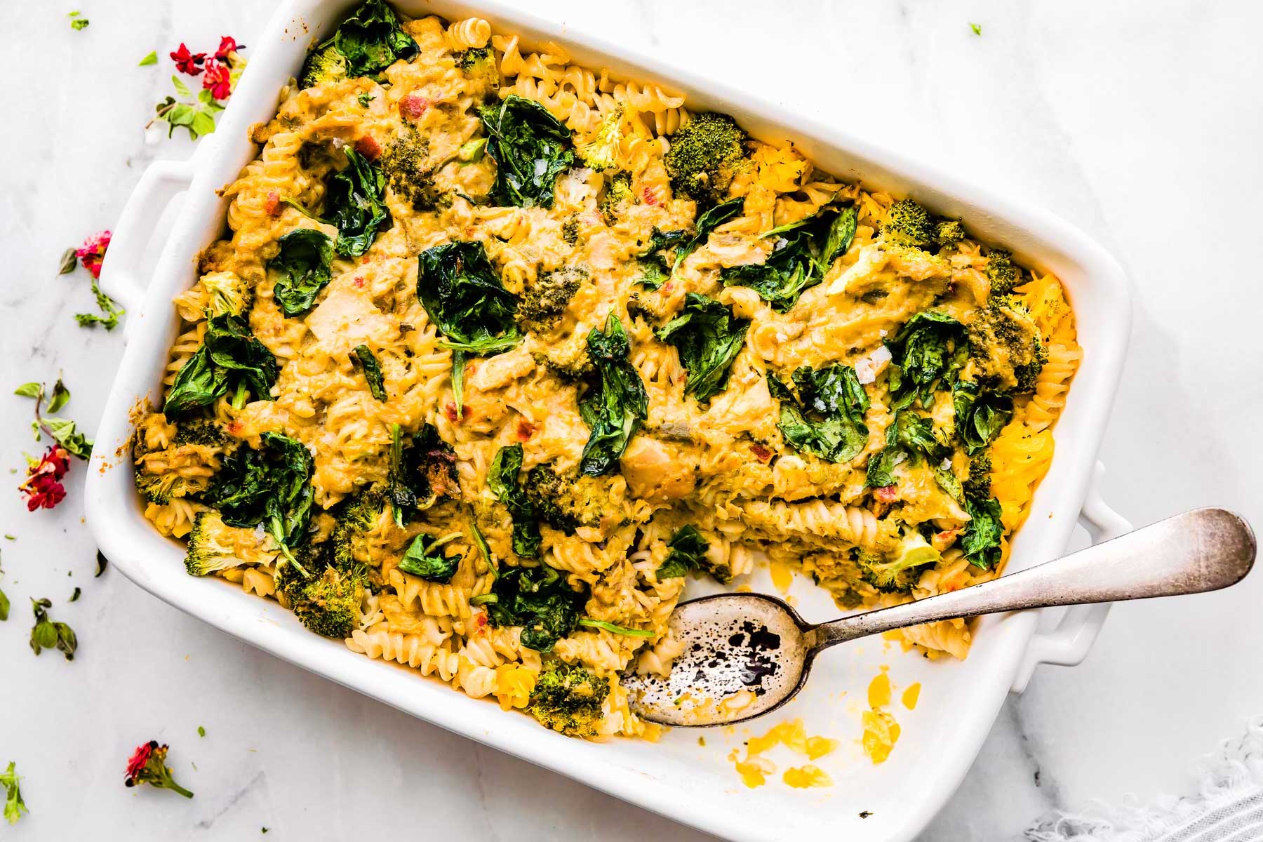 White baking dish with tuna noodle casserole with spinach and broccoli, a serving removed from corner with spoon resting on dish.