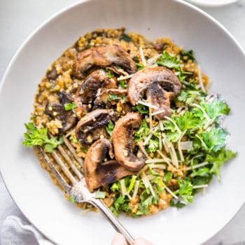 Creamy mushroom risotto served in white bowl topped with mushrooms and fresh greens, a hand holding fork over bowl