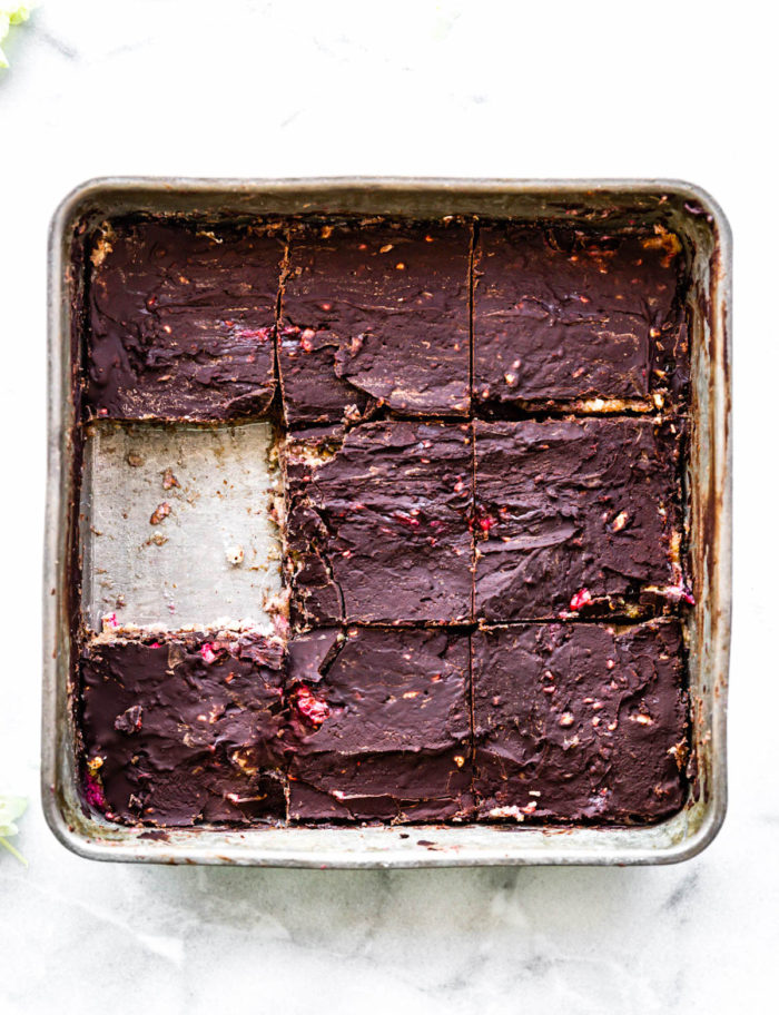 Overhead view silver square pan filled with no bake almond chocolate protein bars topped with chocolate, cut into squares. One square bar removed.