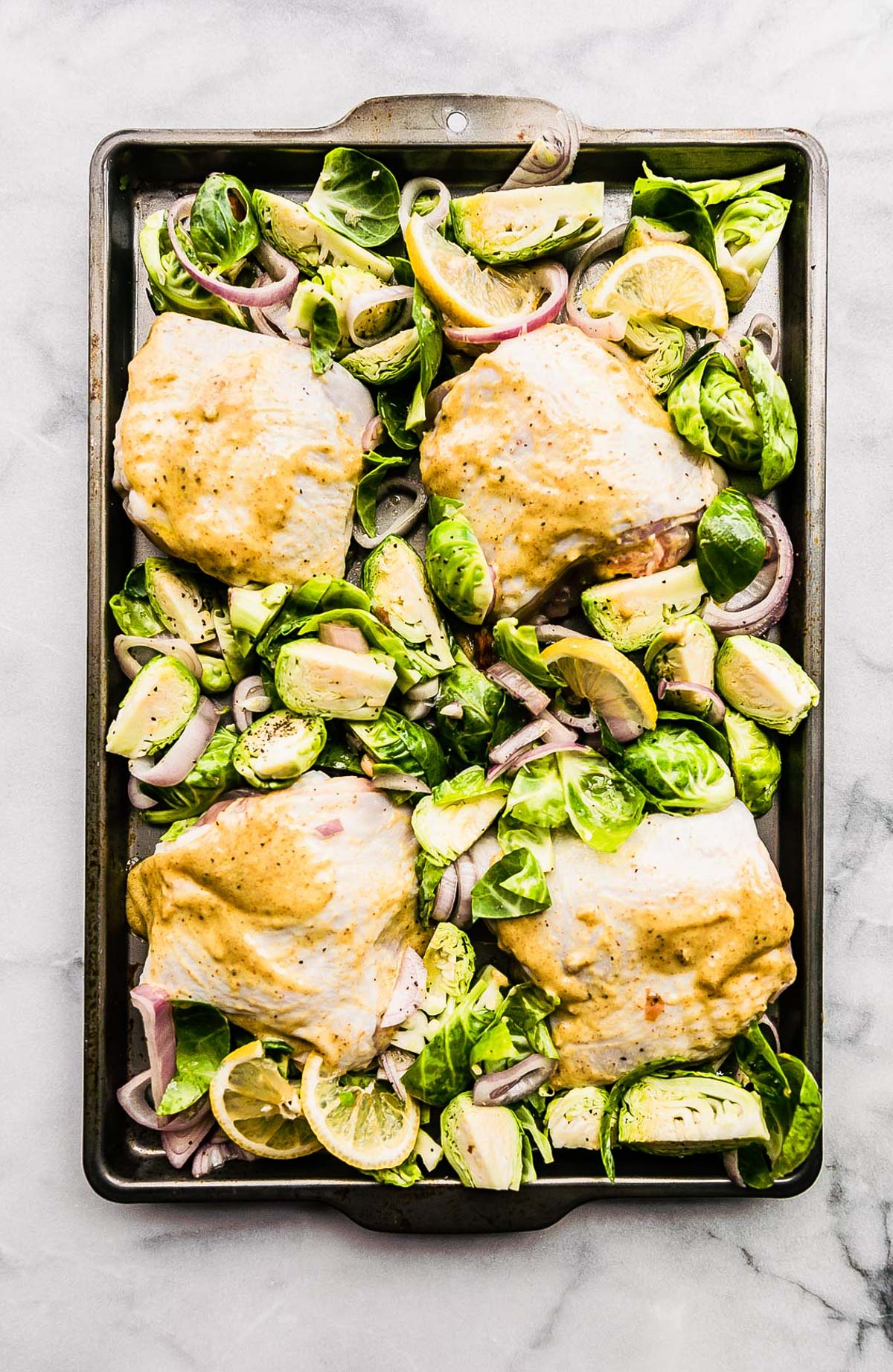 Sheet pan filled with honey mustard raw chicken thighs nestled between sliced Brussels sprouts and red onions