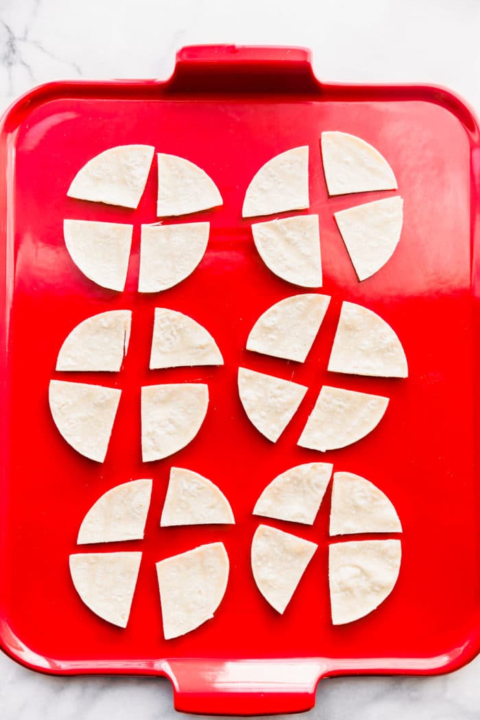Overhead view of six small corn tortillas cut into quarters on a red baking sheet to bake for corn chips to serve with instant pot chicken chili verde.