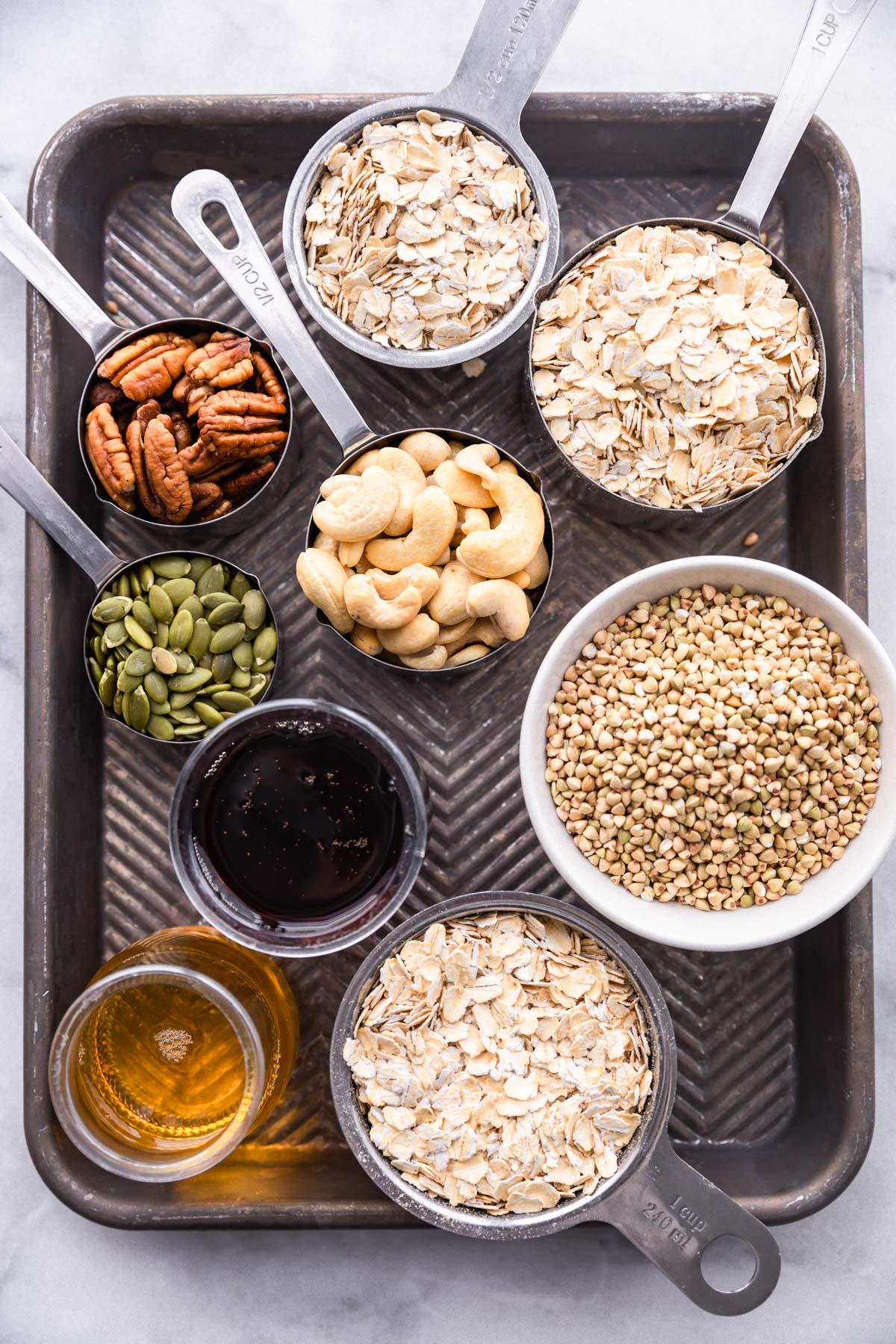 All ingredients for homemade maple buckwheat granola arranged in measuring cups placed on silver baking sheet.
