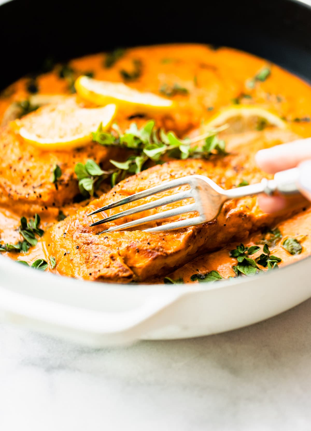 A fork cutting into baked salmon in roasted red pepper sauce still in a skillet.