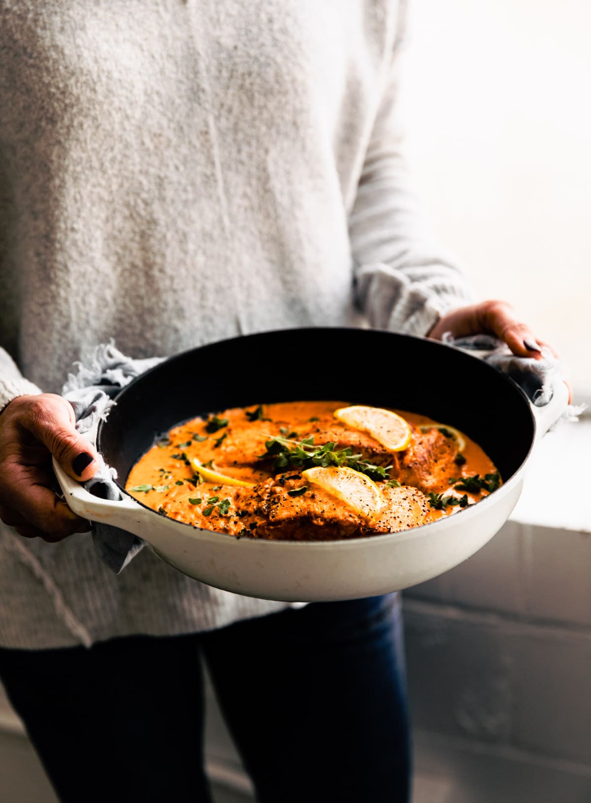 A woman holding a white skillet filled with baked salmon in roasted red pepper sauce.