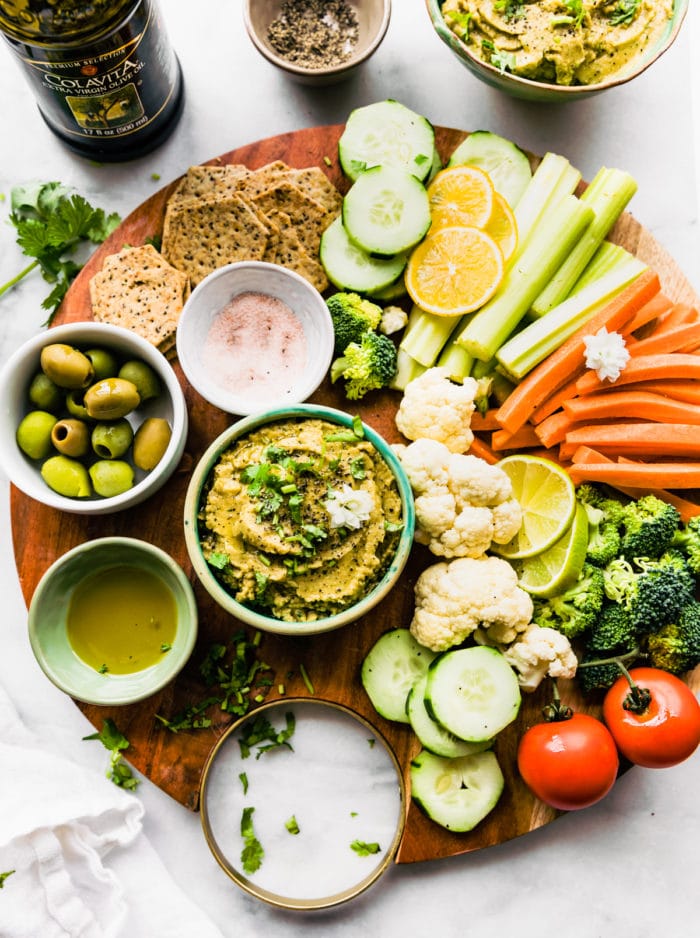 round wooden serving board with assortment of crackers, fresh vegetables, olives, and hummus