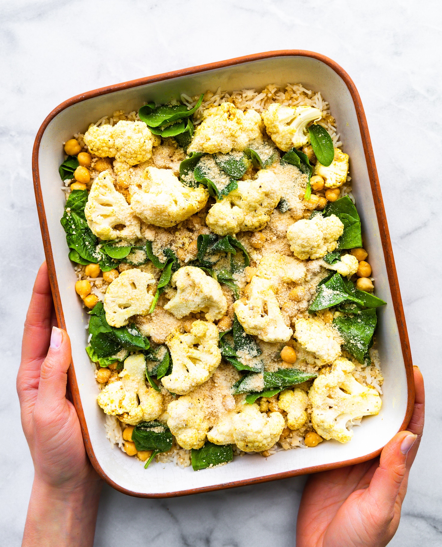 Two hands holding white baking dish at angle filled with curried chickpea cauliflower bake with spinach and nutritional yeast on top.