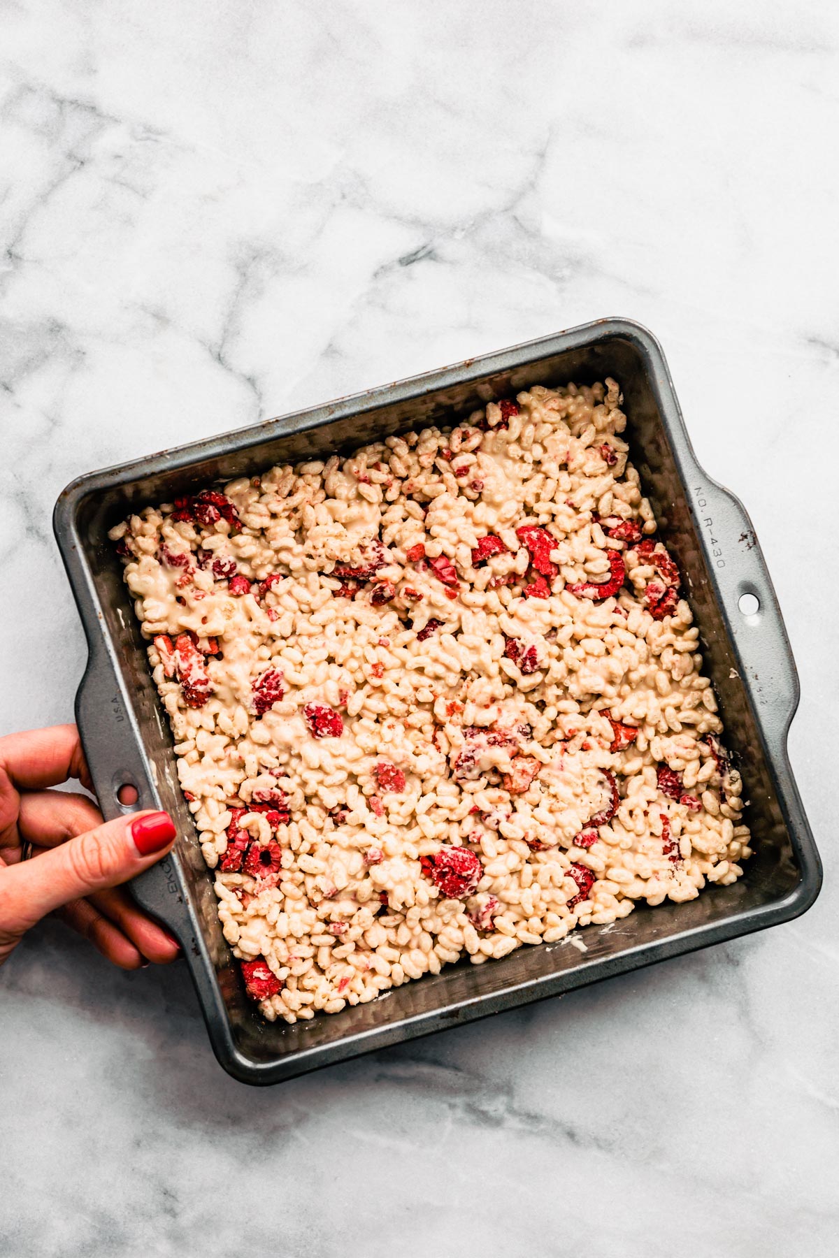 Overhead view a woman's hand holding onto side of baking pan filled with white chocolate rice crispy bars with dried raspberries.