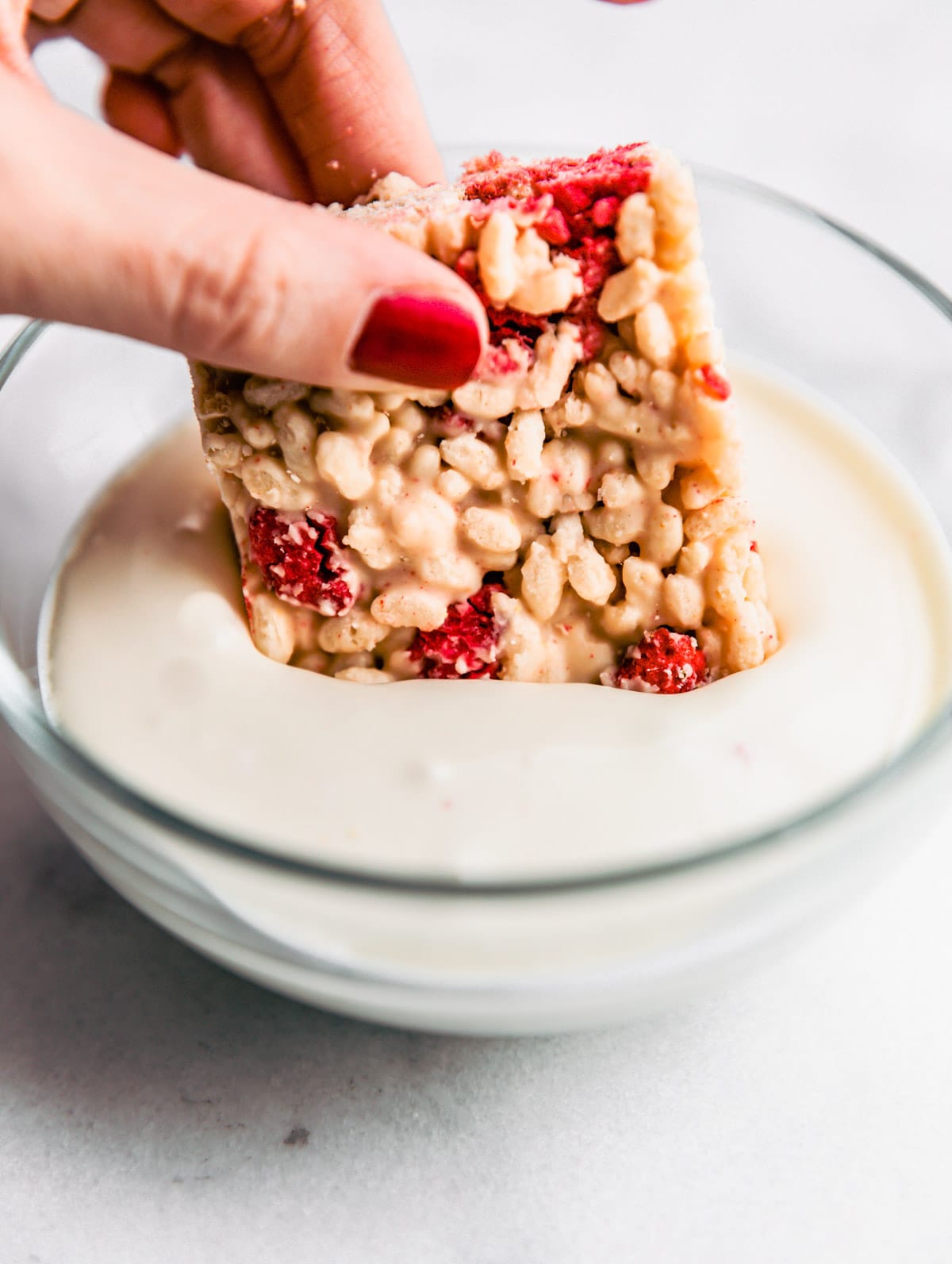 A woman's hand dipping a white chocolate raspberry rice crispy bar into melted chocolate.