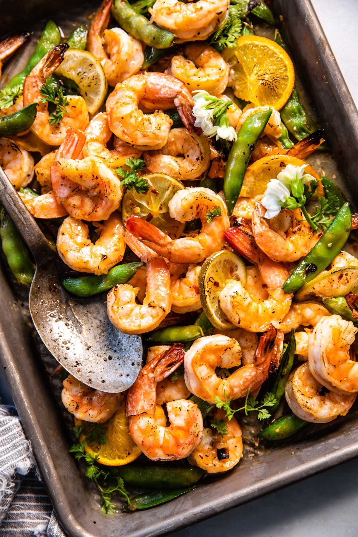 Overhead view sweet and spicy broiled shrimp with super snap peas, orange and lime slices on deep baking dish with large serving spoon.