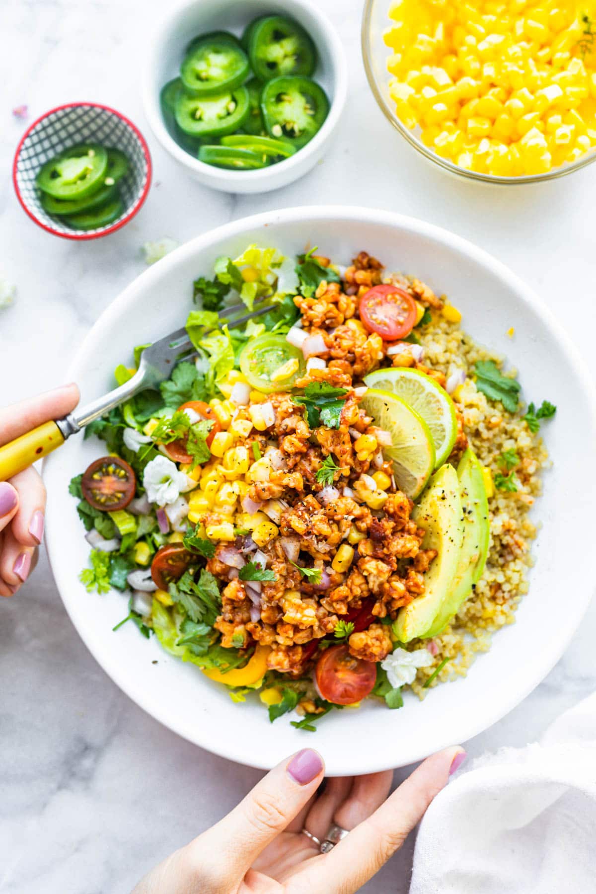 BBQ Tempeh quinoa salad topped with corn, limes, tomatoes, and avocado slices served in white bowl, a hand holding a fork going into salad.