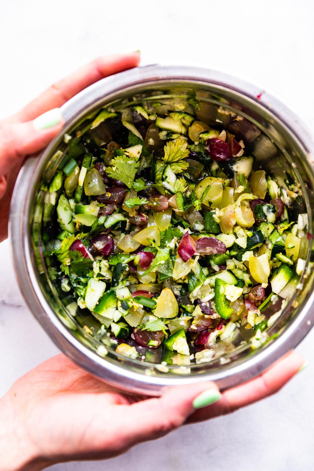 two hands holding bowl of homemade cucumber salsa with grapes and jalapeno