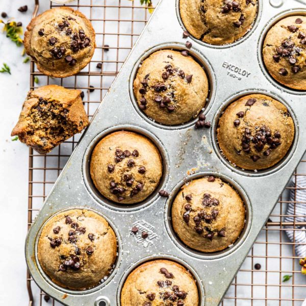 Chocolate chip Muffins in muffin pan. Sitting on cooling wrack. Two muffins out of pan and one with bite taken out. Spoon of sugar above pan