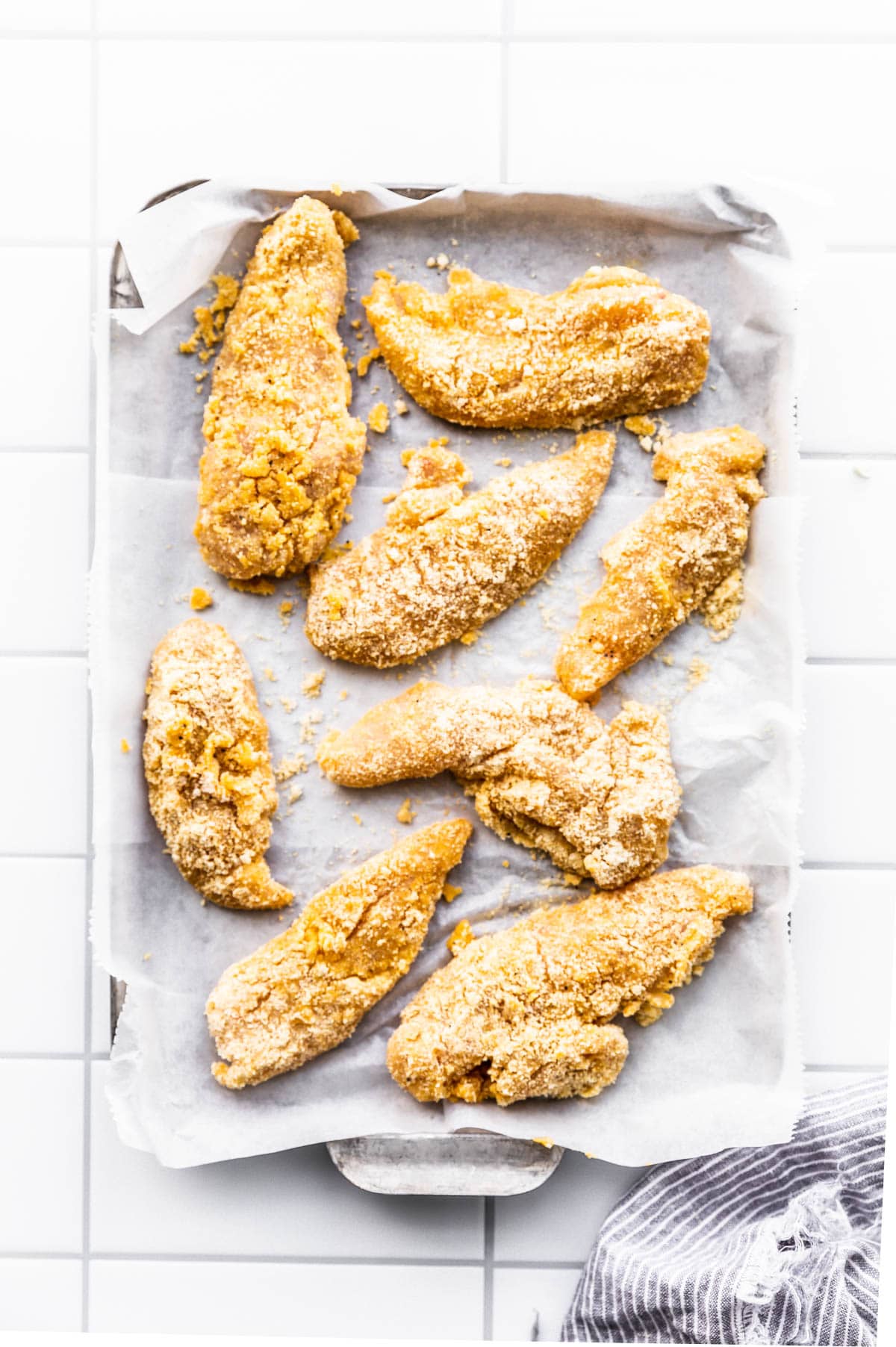 uncooked chicken tenders with breading on spread out on parchment covered baking sheet.