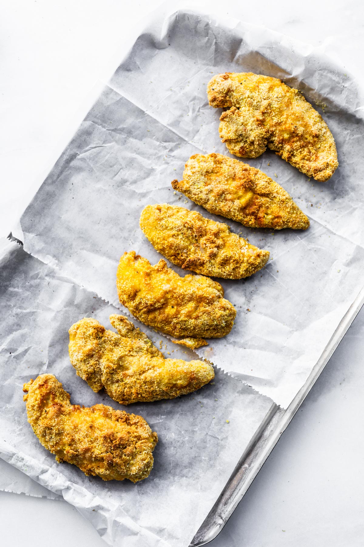 Overhead view parchment covered baking sheet with golden chicken tenders lined up.
