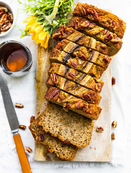 overhead photo: loaf of low carb banana bread cut into slices on wooden board next to knife and honey
