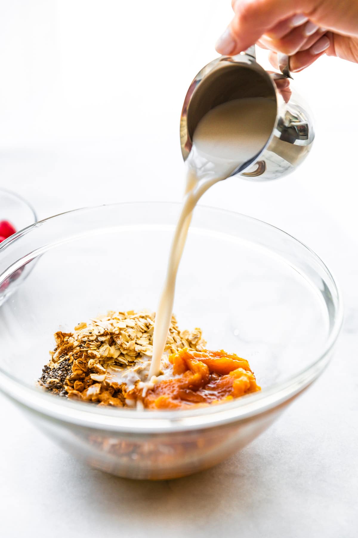 A small silver carafe of condensed milk being poured into a glass mixing bowl filled with the ingredients for vegan pumpkin overnight oats.
