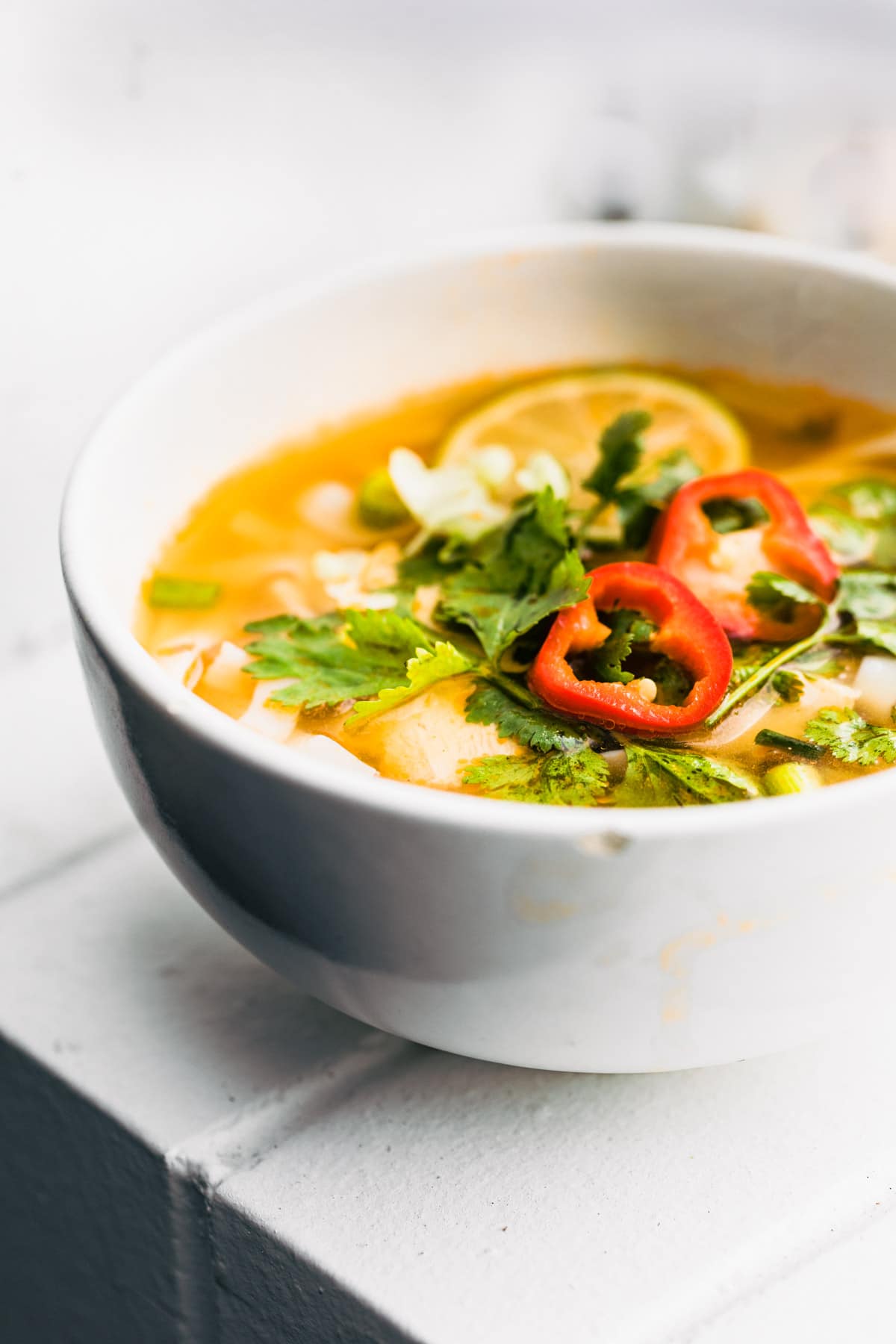 A homemade Thai Chicken Soup recipe in a white bowl on a white countertop.