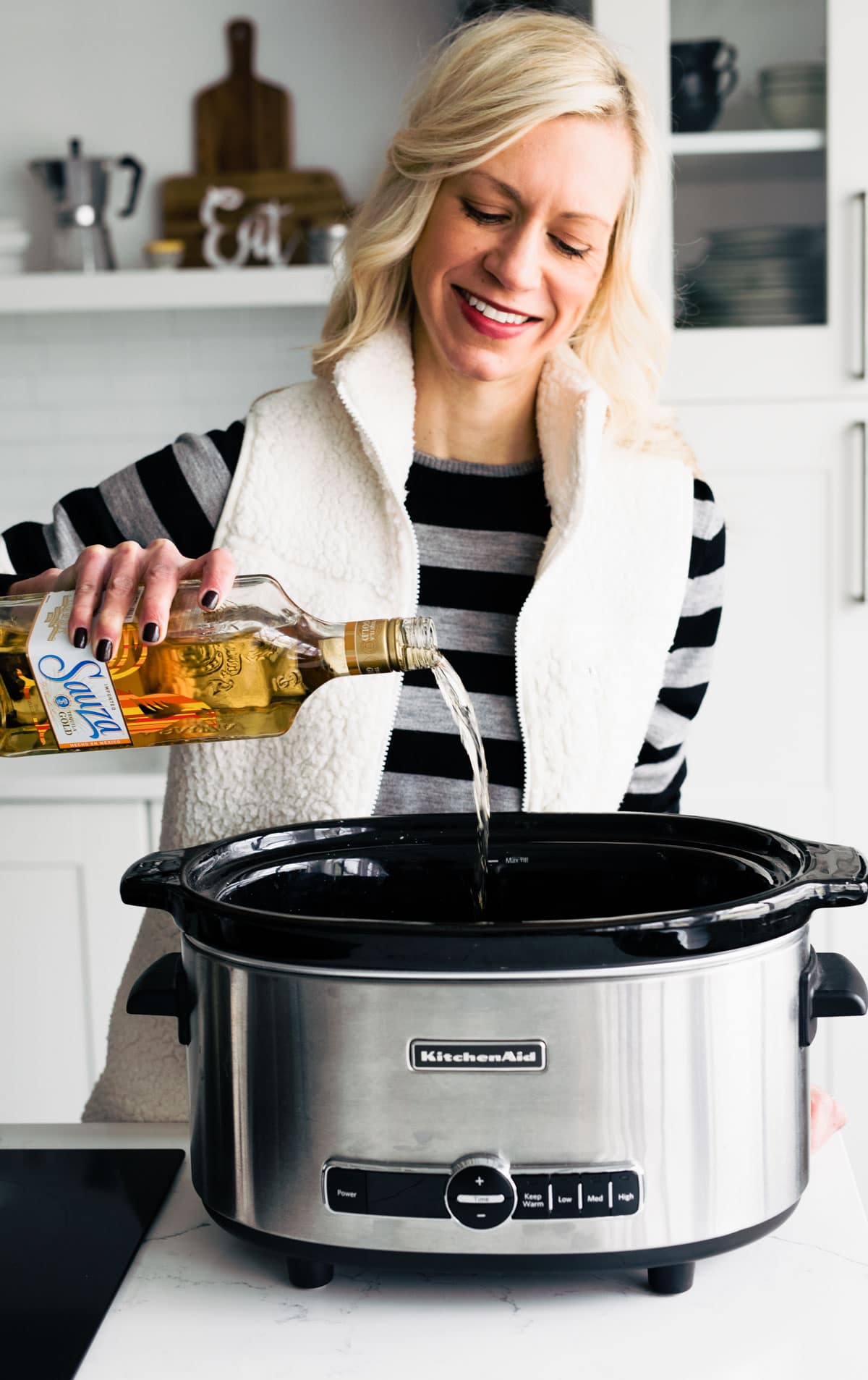 A woman smiling as she pours liqueur into a slow cooker in a white kitchen.